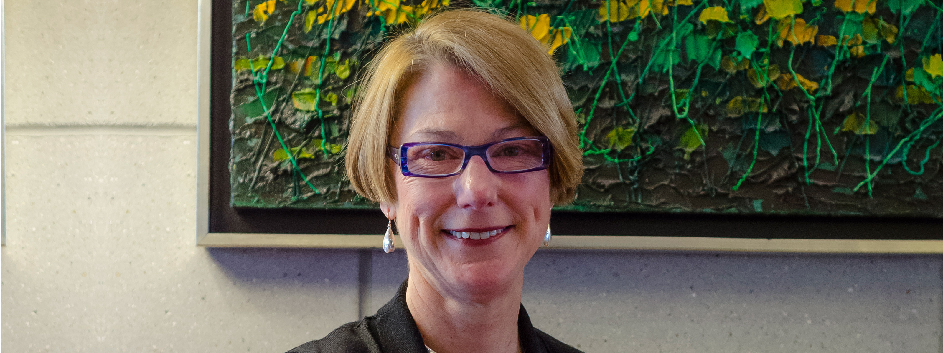 A headshot of a woman smiling at the camera, with a painting of flowers in the background.