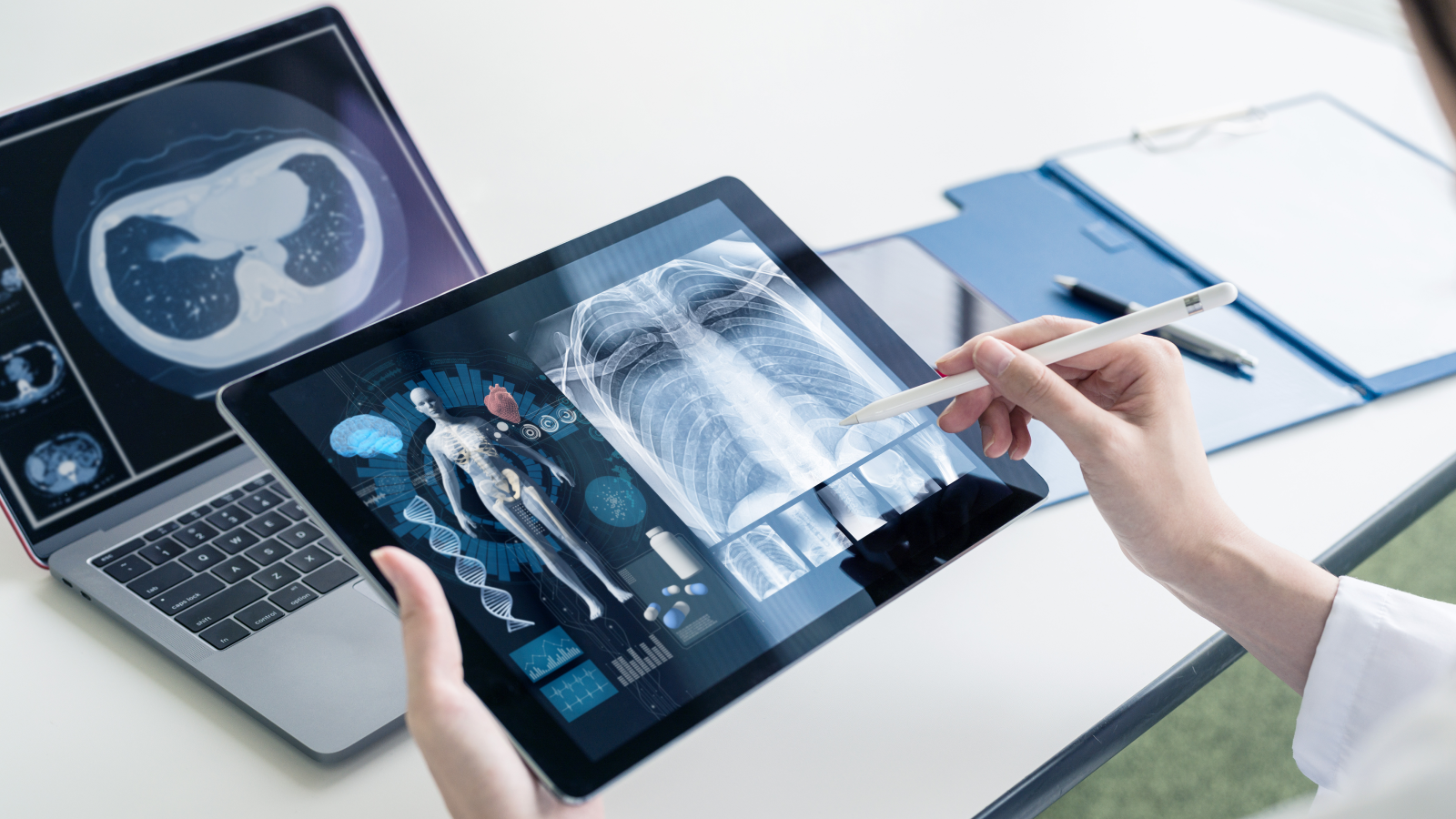 A close-up of someone holding a tablet and stylus with a visual of human anatomy on it; a laptop and notebook are in the background.