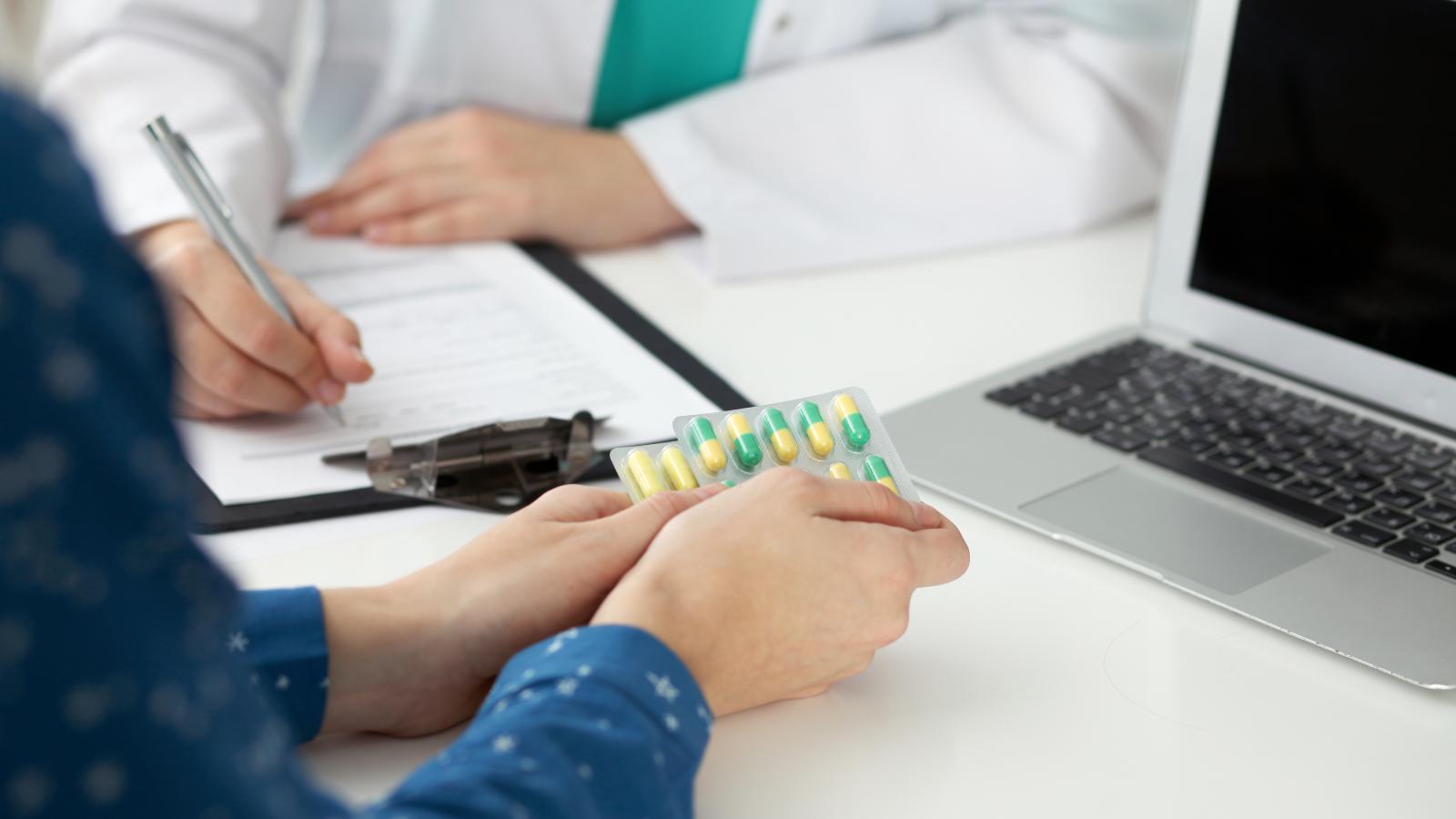 A close-up of two people, one of whom holds a packet of pills, the other of whom is a pharmacist writing a prescription.