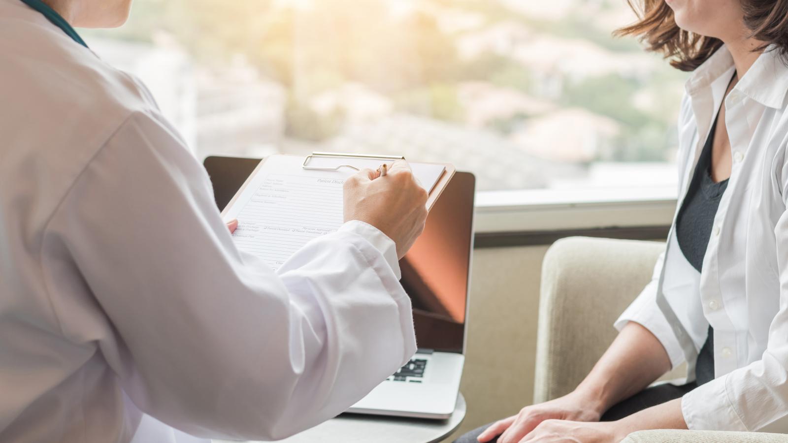 A health care practitioner writing on a clipboard and speaking to a patient.