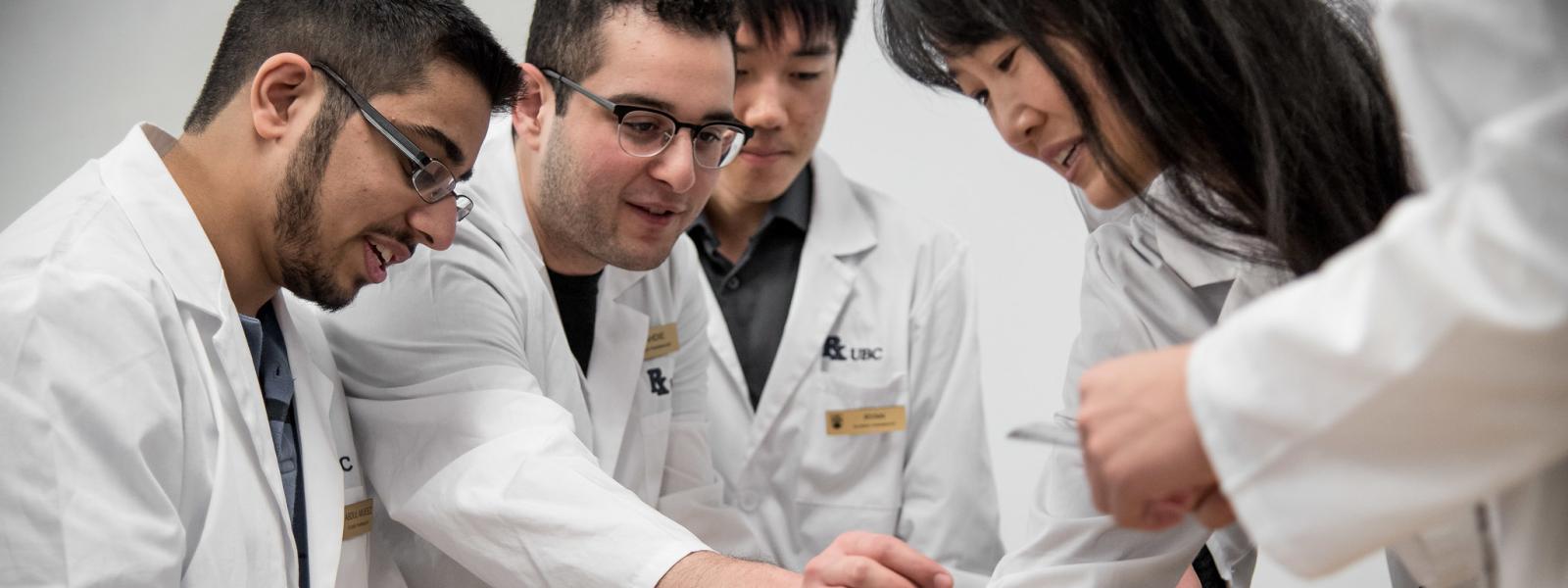 Four students in lab coats speaking and gesturing at something below the bottom edge of the photo.