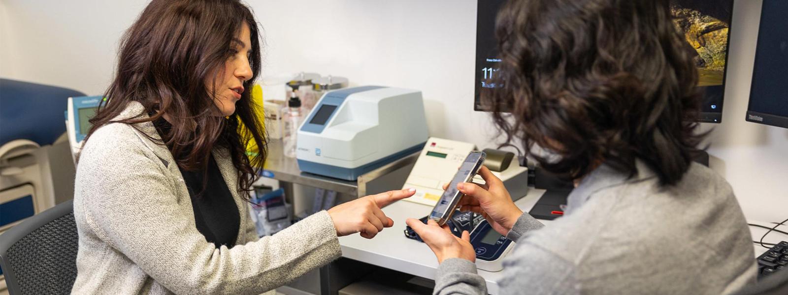 A clinic office where a pharmacist is pointing to a phone which the patient is holding.