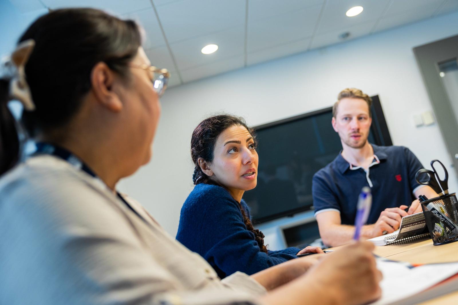 Two women and one man sitting and speaking to others offscreen in a meeting room.