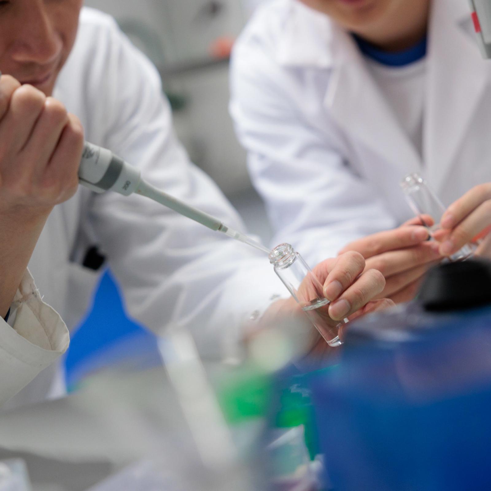 A close-up of two researchers in a lab; one is holding a syringe and they are both holding test tubes.