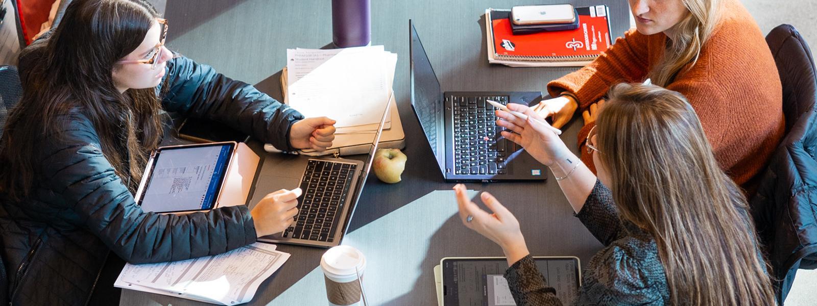 A top-down view of three people conversing at a large desk covered in laptops and notebooks.