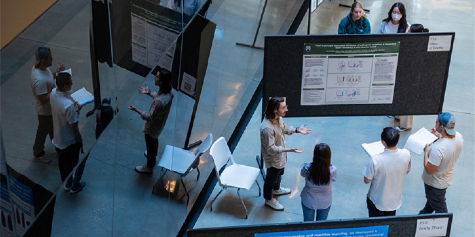 A group of students and researchers gathered around a research poster in a large indoor atrium.