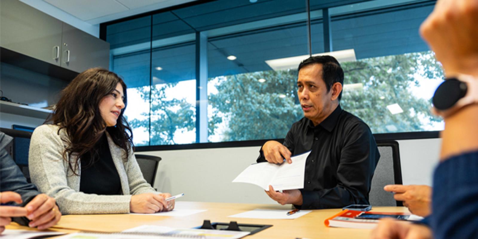 A man gesturing at a brochure while a woman, and others offscreen, look on in a meeting room.