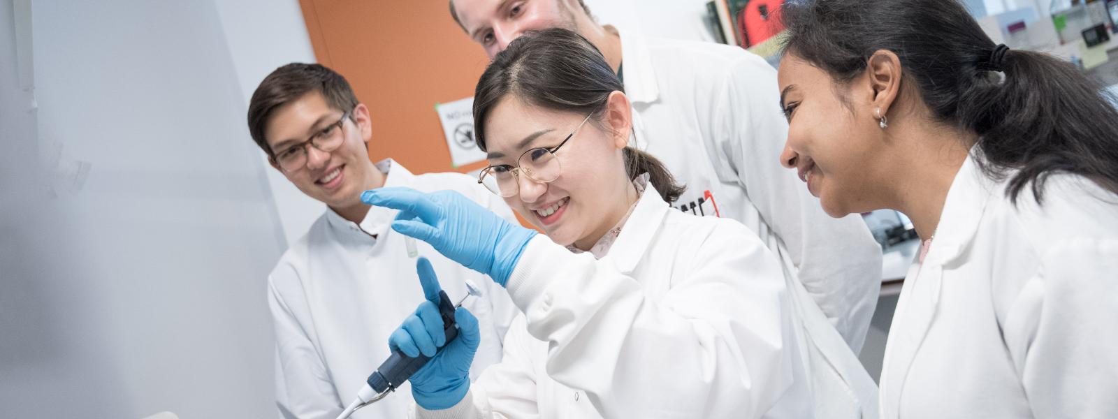 Four students wearing lab coats in a lab; the student in the middle is holding some lab equipment and smiling while the others look on.