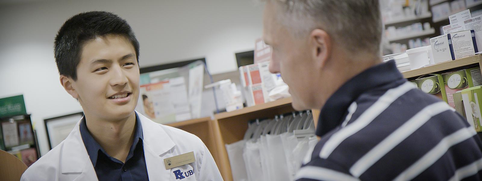 A student in a lab coat speaking to a patient at a pharmacy.