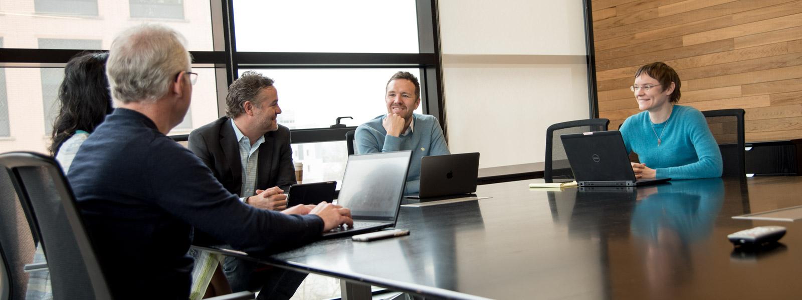 Five people conversing at a large table with laptops, in a spacious, well-lit room.